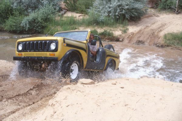 A man driving a yellow and black International Scout II up the steep and sandy bank of a river. The spinning wheels are kicking up water and sand.