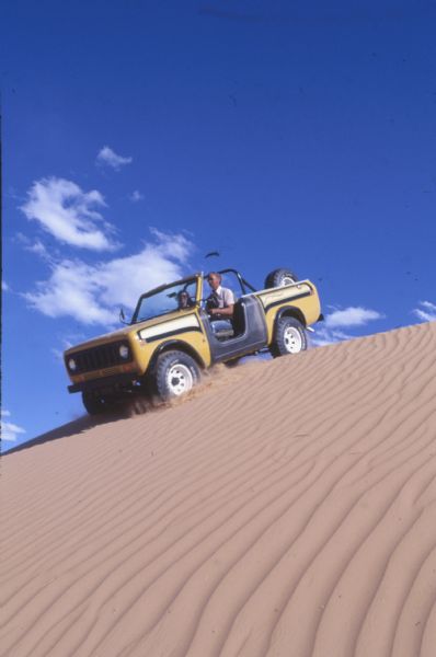 Low angle view up dune towards a man and woman driving a yellow and black International Scout II coming down the side of a sand dune.