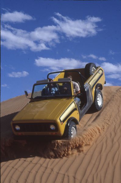 A man and woman are driving a black and yellow International Scout II down a steep sand dune.