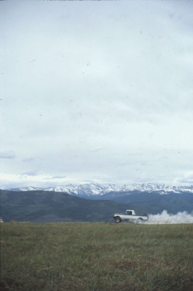 A man driving a white International Scout Terra over a grassy field. There is a large cloud of dust behind the Scout, and rolling green hills and a mountain range in the background.