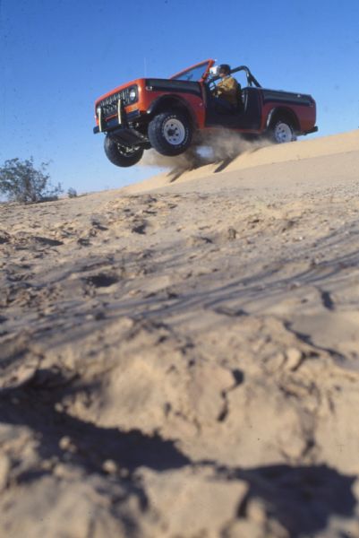 A man driving a red International Scout SS II comes over the crest of a sand dune while off-roading in the desert.