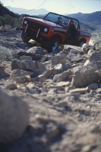 A man drives a red International Scout SS II up a hill in rocky terrain. Mountains are in the far background.