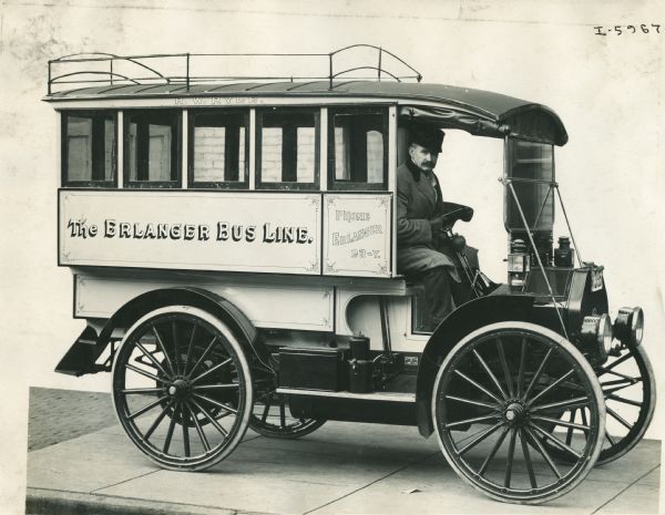 Three-quarter view from front of right side of an International bus used by the Erlanger Bus Line. A man sits behind the wheel of the bus. Above the row of windows a sign is painted that reads: "H.W. Ryle." There is a luggage rack on the roof.