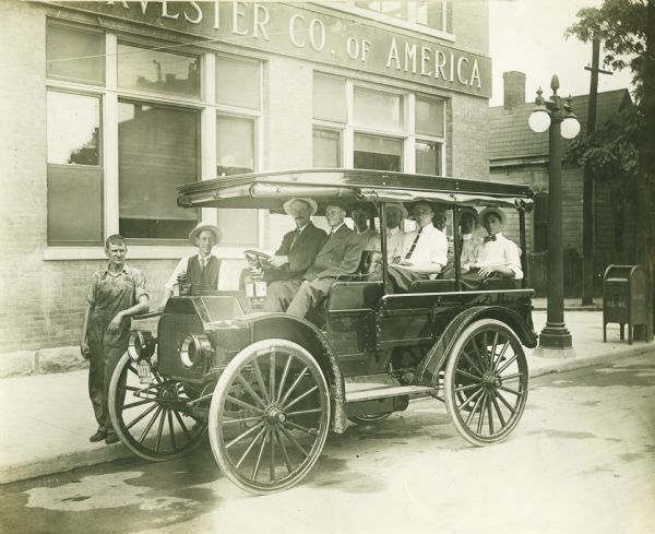 Three-quarter view from front of a group of men sitting in an Internatioal bus which is parked at the curb near what appears to be an International Harvester Company dealership building. Two men stand behind the vehicle on the sidewalk. The side and back flaps are rolled up against the roof, and most of the men are wearing hats and neckties. Behind them is a large lamppost and a U.S. Mail post office box.
