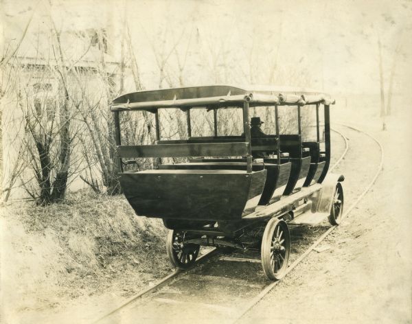 Three-quarter view from rear of the right side of a bus with bench seating operating on metal tracks. The flaps of the bus are rolled up against the roof, and a man wearing a hat sits in the driver's seat. On the left is a building behind trees and bushes. The bus is probably built on an International truck chassis.