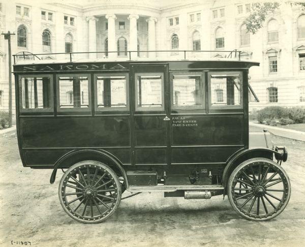 Side view of right side of an International bus marked "Nakoma" parked in front of the Wisconsin State Capitol. The bus has an enclosed cab and the passenger door has a painted sign near the handle that reads: "Pay as you enter Fare 5 cents." There is a luggage rack on the roof.