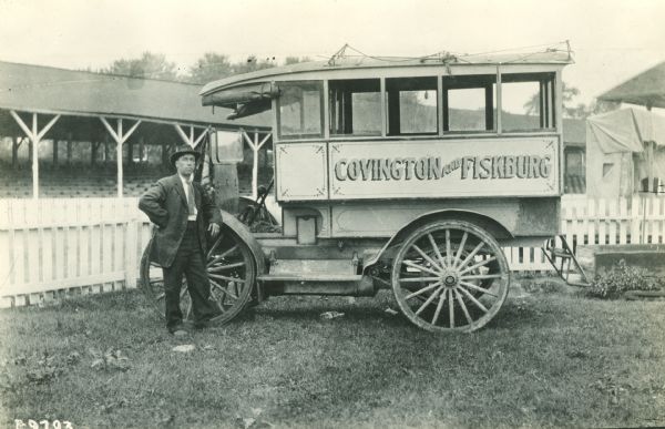 A man stands on a lawn by the front wheel of an International bus marked "Covington and Fiskburg." In the background is a fence and beyond that is a covered seating area.
