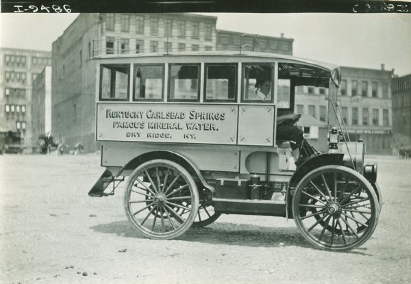 Side view of right side of bus marked: "Kentucky Carlsbad Springs Famous Mineral Water. Dry Ridge, KY." A man sits in the driver's seat of the open front cab. In the background are large brick buildings.
