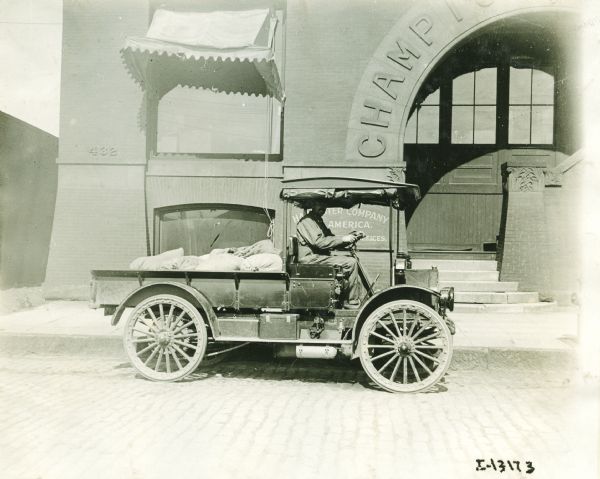 A truck parked in front of an International Harvester Company building, possibly the Champion Works. The building has an archway, with lettering which reads, in part: "CHAMPI ..." A man sits in the driver's seat of the cab, which has the side and back flaps rolled up to the roof. Filled burlap bags are stacked in the open bed.