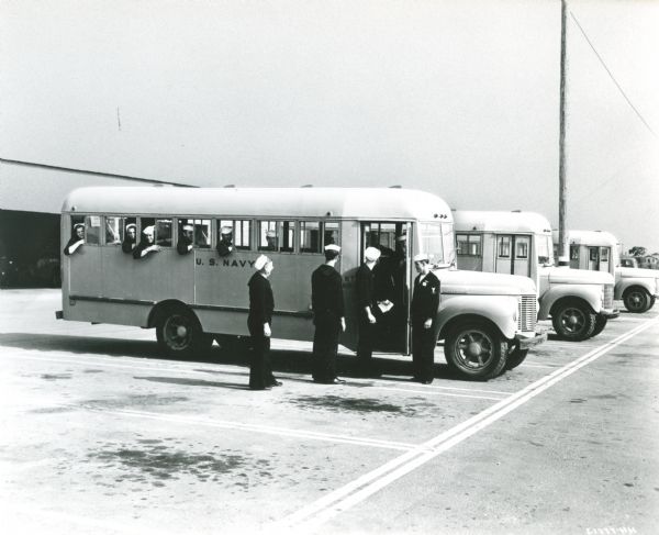 Men in Navy uniforms enter an International bus marked "U.S. Navy." Men sitting in the bus are leaning out the windows on the right side. There is a long building, perhaps a garage, in the background, and more buses are in the background in parking spaces.