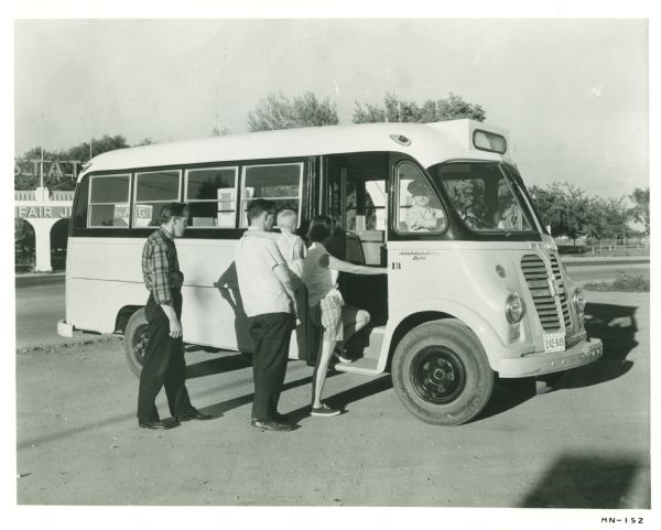 Four people board an International Metro school bus. A man wearing a hat sits in the driver's seat. A sign behind the bus reads: "State Fair."