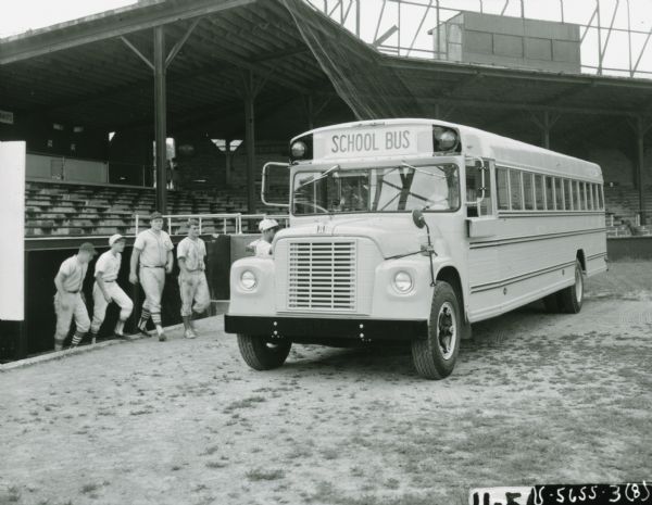 Men and boys in baseball uniforms walk out of a dugout to board a Wayne school bus parked inside a baseball stadium.