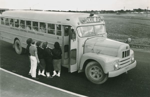 Slightly elevated view of a group of four young women and one young man as they board an International R-line school bus parked along a rural road. The text on the side of the bus reads, "Gates - Chili Central School Operator."