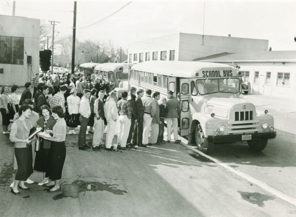 A large group of school children, probably high school age, are standing in line to board an International R-line school bus. Two more buses are in the background.