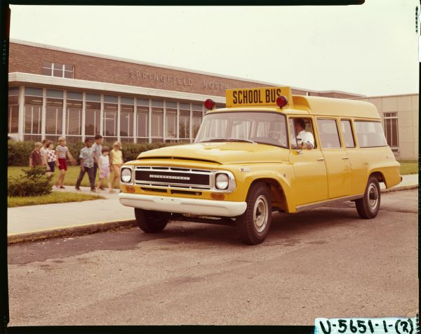 Young children are walking down a sidewalk to board an International C-1200 school bus parked outside Springfield North School. A man is sitting in the driver's seat.