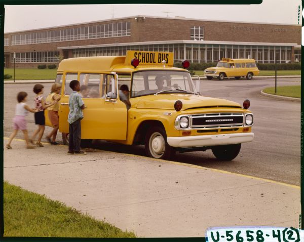View towards a group of students boarding an International C-1100 school bus outside Springfield North School. A manis  sitting in the driver's seat.