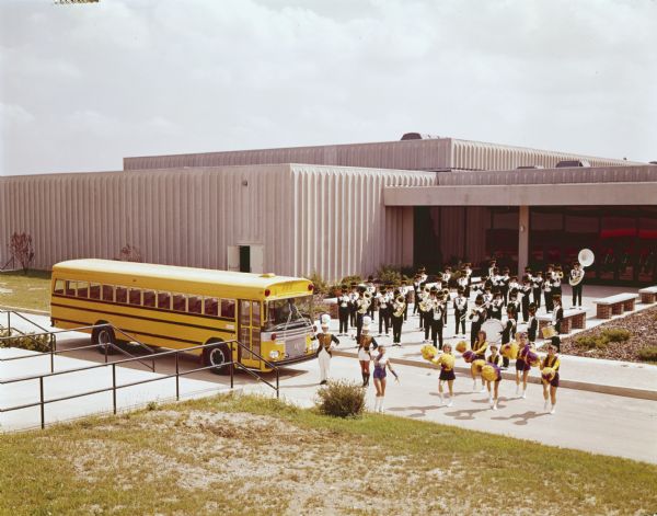 View looking down hill towards an International school bus with a Wayne body parked outside a building, surrounded by a marching band posing on the front steps, and a baton twirler and cheerleaders on the road in front of the bus.