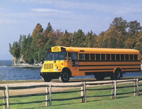 Color photograph of an International 3600 Vista school bus on a gravel road along a shoreline. There is a man in the driver's seat. In the foreground is a rustic fence and lawn, and in the background is a rocky shoreline with trees.