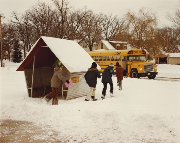 Color photograph of kids walking through snow while leaving a metal bus shelter to board an International school bus. There are houses in the background.