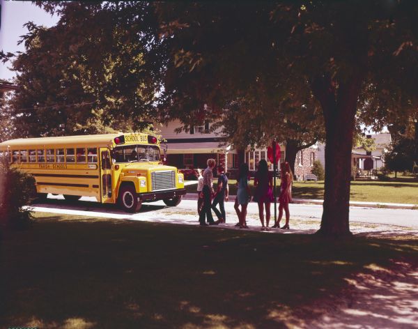 Color photograph of a group of children standing on a sidewalk to wait for an approaching school bus marked "Parish Schools." The bus is built on an International Loadstar 1700 chassis with a body by Carpenter.