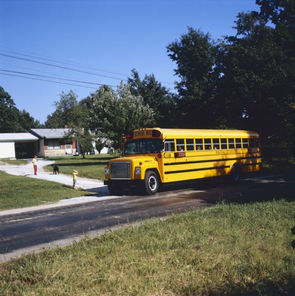 Color photograph of two children and a dog standing in the driveway of a residence while an International bus with Carpenter body passes by. The side of the bus is marked: "Parish Schools."