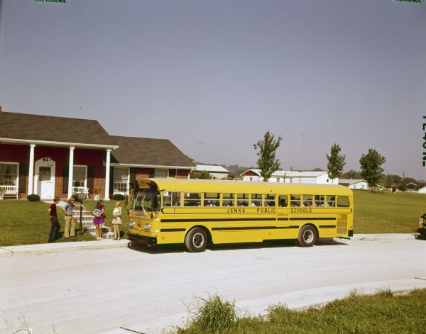 Color photograph of four children holding books and standing beside a gravel road in front of a suburban residence. An International rear-engine drive school bus owned by Jenks Public Schools has pulled up to the driveway.