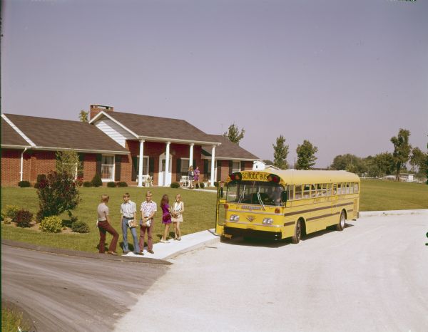 Color photograph of a group of teenagers holding books and standing on the sidewalk in front of a suburban residence. In the background two girls are walking near the porch towards an International rear-engine drive school bus parked at the curb with the door open.