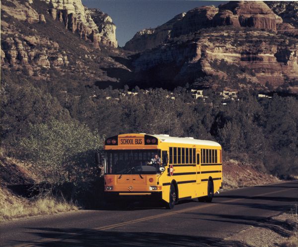 Color photo of a person driving an International Harvester school bus. This photograph was likely taken for promotional or advertisement material for the bus line. On the low hills behind the bus are low, adobe-style buildings hidden partially behind trees. In the far background is a desert mountain landscape.