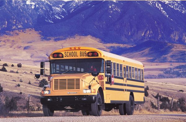 Color photo of a person driving an International school bus in the southwest United States. In the background is a snow-capped mountain range. This photograph was likely taken for promotional or advertisement materials.