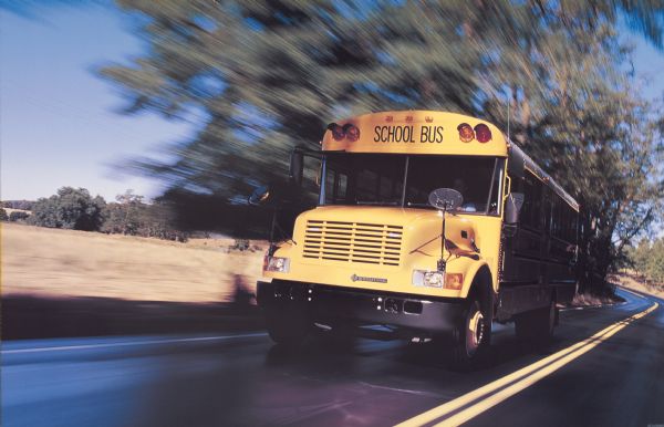 Color photo of an International school bus, shown in a blur of motion on a road. The "International" emblem is on the grille of the bus, and there is a male bus driver. In the background are trees and open space. This image may have been used for advertisement materials.