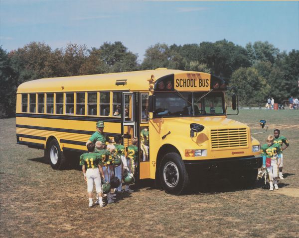 Color photo of an International school bus parked on a football field. Several youth football players and a coach are dressed in white, yellow, and green football uniforms, and they are filing onto the bus. In the background is a group of adults and practice equipment. Below the grill of the bus is the "International" emblem in red. This photograph for was probably taken for advertisement materials.