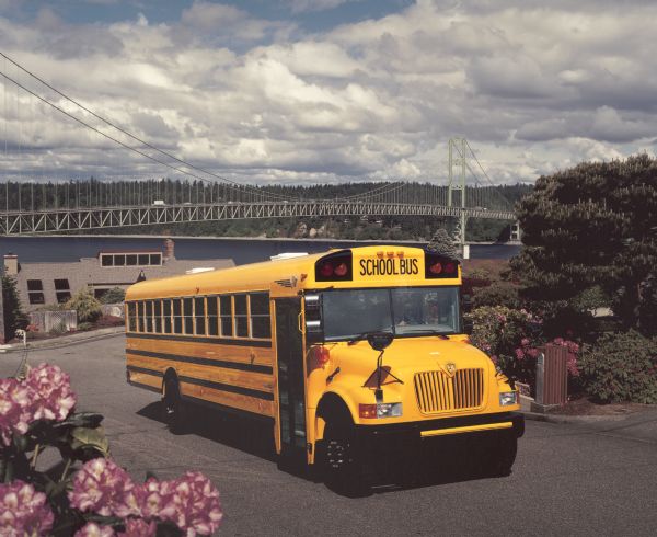 Color photo of an International school bus with a man in the driver's seat. The bus is in the middle of a street in a hilly residential area, with landscaping of trees, flowers, shrubs and fences. In the background is traffic on a large bridge over a body of water, with a forested shoreline beyond. This photograph was probably taken for advertisement materials.