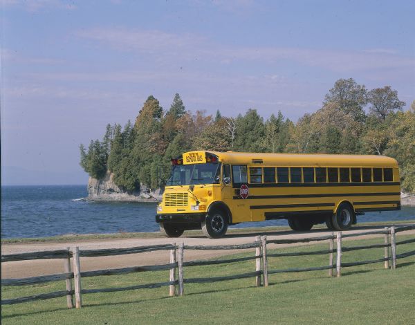 Color photo of an International school bus driven along a coastal road. In the foreground is a split-rail fence and lawn. The photograph was probably taken for advertisement materials.