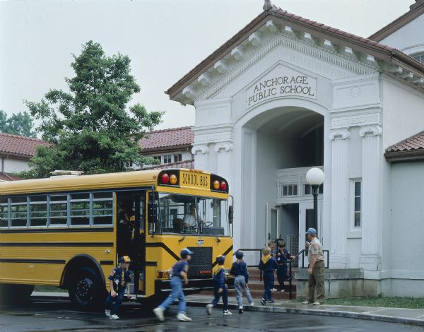 Color photo of a group of Cub Scouts and their scout leader exiting an International school bus and entering the Anchorage Public School. A woman is driving the bus. This photograph was probably taken for advertising materials.