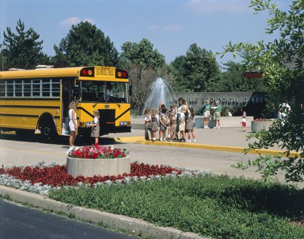 Group of Brownie Girl Scouts and their leader are posing next to an International school bus at the Louisville zoo. In the background zookeepers and children are near the front of the building. A woman is driving the bus. This photograph was probably taken for advertisement materials.