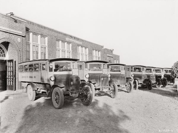 A fleet of International Harvester school buses parked in a row in front of a brick building. Each school bus is equipped with a stop sign and a driver. The bus on the far left has the words: "Jefferson TP. Schools No. 1." painted on the side. A number of men are sitting inside the bus.
