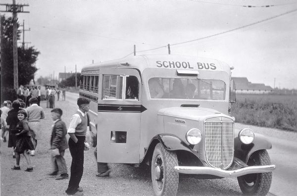 View down side of road of a group of children exiting an International school bus. On the front and side of the bus is painted the "International" emblem. In the background a group of older children or adults are watching the younger children.