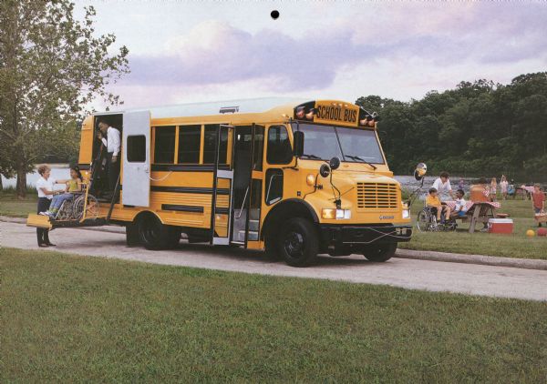 Color photo of a man and a woman assisting a girl on a wheelchair lift for a model 3600 Special Needs bus. The "International" emblem is on the front of the bus. In the background, children and adults are picnicking at a park near a lake or pond. The caption on the photograph reads: "International 3600 Special Needs Bus with Thomas Built Vista Body".