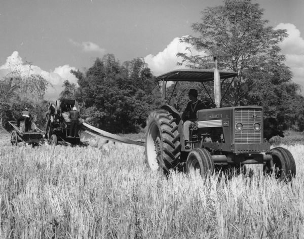 This photograph was likely taken by the International Harvester Overseas Division for promotional purposes, and shows a man driving an International Harvester 624 tractor while two other men work a rice thresher. The men are dressed in pants, long sleeve shirts, and hats. The tractor model is on the front of the tractor and has the "McCormick" and "IH" insignia. The original caption reads: "McCormick International 624 Diesel Tractors in wetland preparation for rice culture, unhusked rice (palay) threshing, and sugarland preparation. Scenes such as those shown in the accompanying pictures are common in the Philippines, an agricultural country, whose principal crops include rice and sugar."