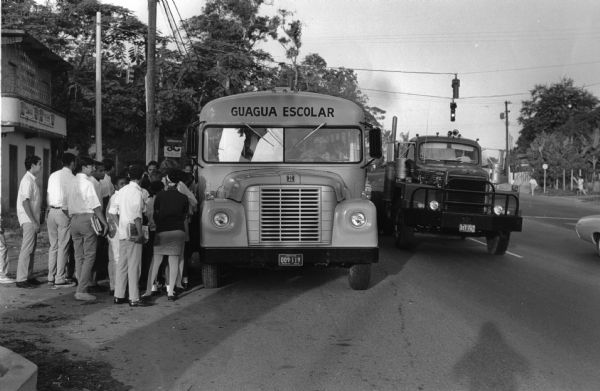 This photograph was likely taken by the International Harvester Overseas Division for promotional purposes. It shows students wearing school uniforms and holding books while waiting to board a model 1603 60 passenger school bus. The "IH" insignia is just above the grille of the bus and the top of the bus reads: "GUAGUA ESCOLAR". The original caption to the photograph reads: "This bus is picking up school children on rural acres as per a new program started by the Commonwealth of P.R. 2 years ago and in which they supply free of charge, transportation for school children living on the rural acres. Because the condition of our rural roads International has been chosen by most of the municipalities because of their sturdiness in construction(?) and at the same time smooth performance." This photograph was taken in the municipality of Toa Baja, Barrio Candelaria, Puerto Rico.