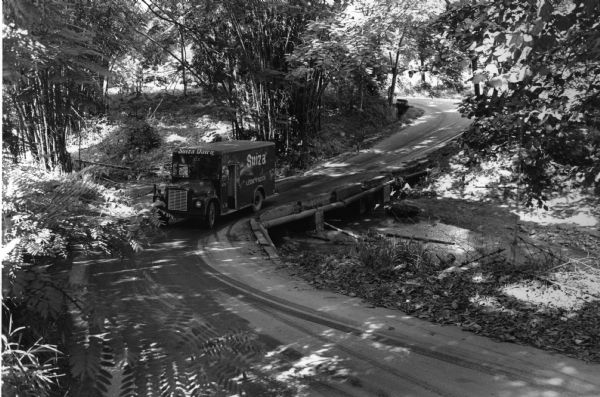 This photograph was likely taken by the International Harvester Overseas Division for promotional purposes, and shows a "Suiza Dairy" International Harvester 1600 chassis truck with refrigerated milk body driving down a forested road. The side of the truck reads: "Suiza Leche Fresca." The "IH" insignia is just above the grille on the front of the truck. The original caption to the photograph reads: "Performance very good 'Surprised' Mr. Felix Romain-Supervisor." The driver of the truck is Marian Nenitez, and the photograph was taken at San Lorenzo Rivera, San Juan, Puerto Rico.