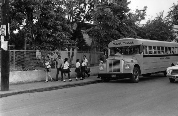 This photograph was likely taken by the International Harvester Overseas Division for promotional purposes, and shows students in school uniforms with books exiting a model 1603 60 passenger school bus. The "IH" insignia is just above the grille of the bus and the top of the bus reads: "GUAGUA ESCOLAR." The side of the bus reads: "MUNICIPIO DE TOA BAJA." The school bus was purchased by the municipality of Toa Baja for a program initiated by the Commonwealth of Purto Rico to supply children living in rural areas transportation to school. This photograph was taken in the municipality of Toa Baja, Barrio Candelaria, Puerto Rico.