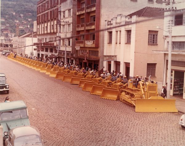 This photograph was likely taken by the International Harvester Overseas Division for promotional purposes. This photograph shows a long line of International Harvester BTD-20 crawler construction equipment on a cobblestone city street. Each tractor has a man sitting on it wearing an "IH" helmet. In the background are other people looking at the tractors on the sidewalk along a row of shops. At the end of the street is a steep hill dotted with homes. This photograph was taken in Joacaba, in the State of Santa Catarina, Brazil, by the International Harvester Overseas Division to mark the occasion of the large delivery to Brazil.