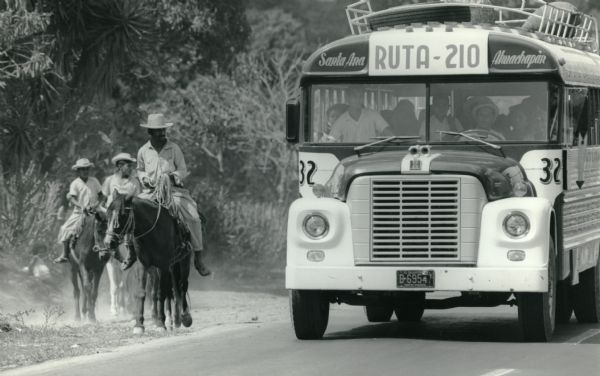 This photograph was likely taken by the International Harvester Overseas Division for promotional purposes. The original caption to the photograph reads: "Bus passengers, with farm produce in rack on top, are transported by IH bus on 21-mile trip between Santa Ana and Ahuachapan, El Salvador." Beside the bus is a man and two boys riding ponies. The IH insignia is on the front of the bus above the grille.