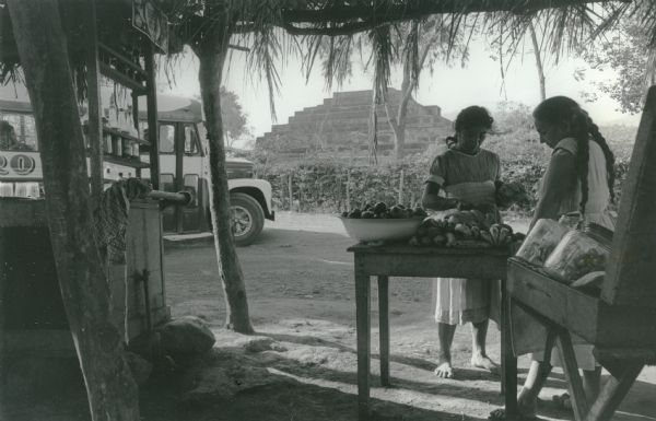 This photograph was likely taken by the International Harvester Overseas Division for promotional purposes. The original caption to the photograph reads: "International bus stops at market place in El Salvador." The photograph shows two women looking at fruit. Around them are wooden tables and shelves filled with glass containers. In the background is an indigenous pyramid.