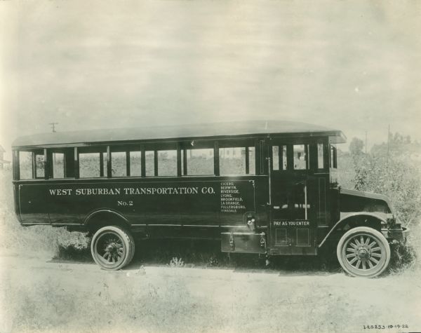 Right side view of a circa 1922 International Harvester bus. The side of the bus reads: "West Suburban Transportation Co. Cicero, Berwyn, Riverside, Lyons, Brookfield, La Grange, Fullersburg, Hinsdale. No. 2". There appears to be a toolbox mounted to the bus just to the left of the passenger door. The passenger door reads: "Pay as you enter". The bus is parked in what is likely the western suburbs of Chicago. Behind the bus are buildings and fields.