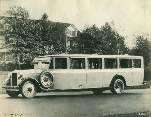 Three-quarter left side view of what is probably a 1924 International Harvester bus parked on a street. The spare tire is mounted on the left side just in front of the driver's side door. In the background is a large house or building. This photograph was most likely taken to be used as advertising and/or promotion material for International Harvester.
