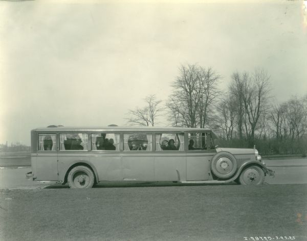 View of right side of what is probably a 1925 International Harvester bus. Passengers, mainly women, can be seen sitting inside, looking out from the curtained windows. A spare tire is mounted near the hood in front of the passenger door. This photograph was most likely taken to be used as advertising and/or promotion material for International Harvester.