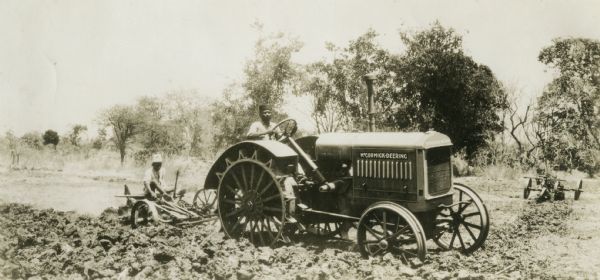 Field scene of a McCormick-Deering 15-30 tractor in India. Shows a  man driving the tractor and one man riding on an implement attached to the tractor. Original caption states: "15-30 plowing owned by FR. [last name unreadable], Dhundesa, India."