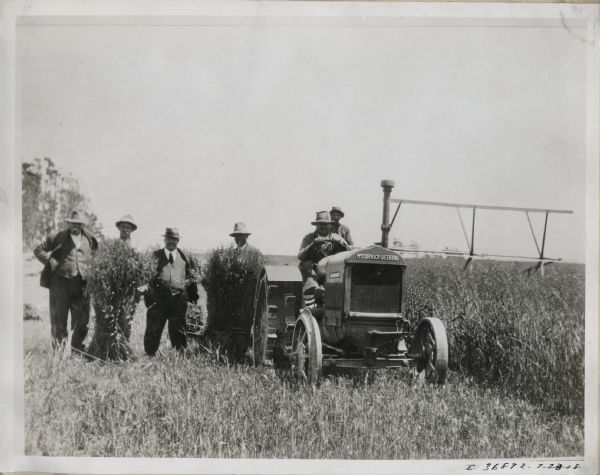 A McCormick-Deering 15-30 tractor in a New Zealand field. Five men stand in a field gathered around bushels of wheat, and one man drives a tractor pulling a implement. Original caption reads: "New Zealand."